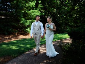 A couple in wedding attire walks along a shaded garden path surrounded by lush greenery.