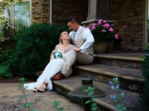 Couple in white clothing poses romantically on stone steps with pink flowers in garden setting.
