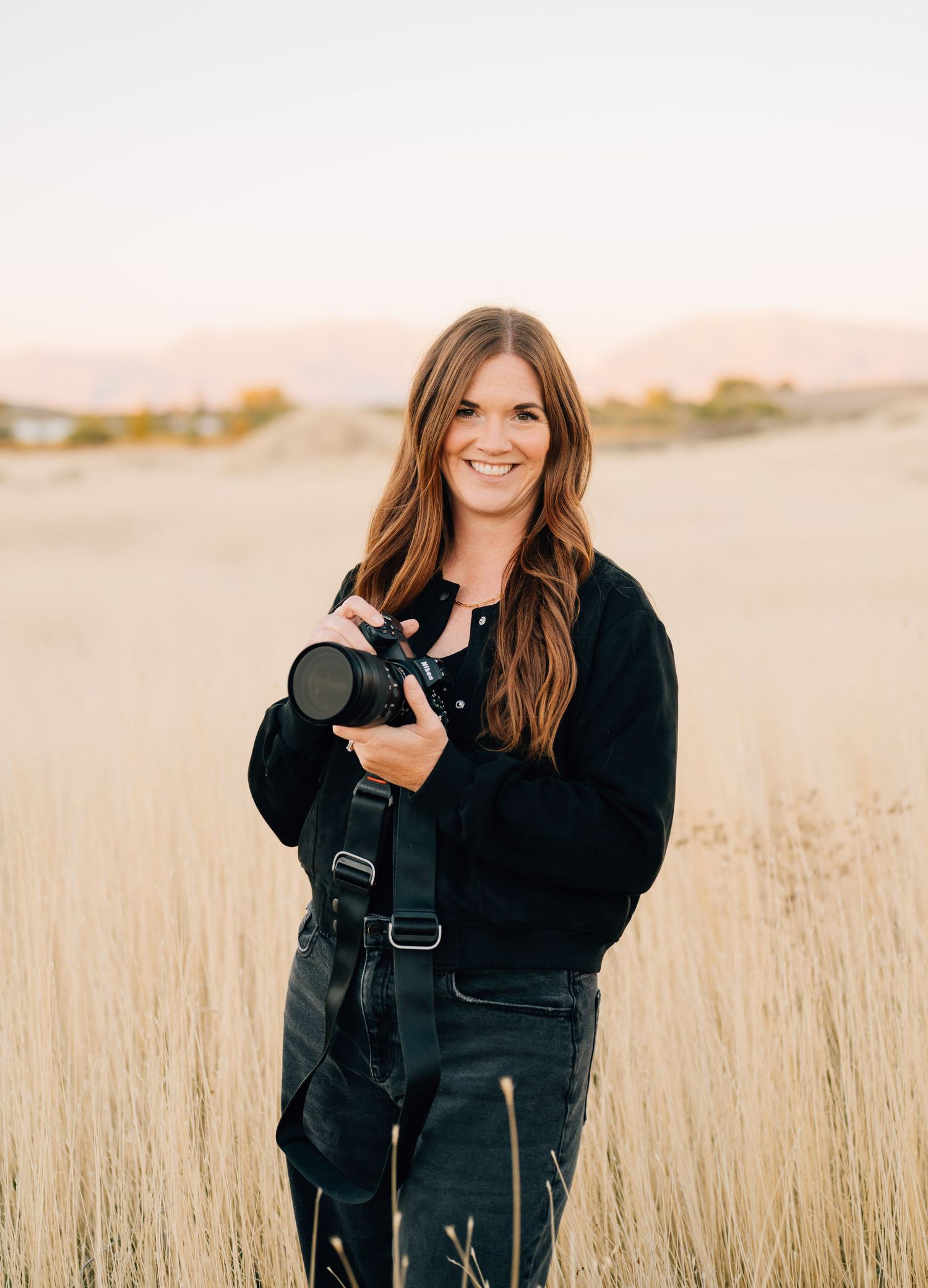 A photographer in a black jacket stands in a golden wheat field during sunset, holding a professional camera.