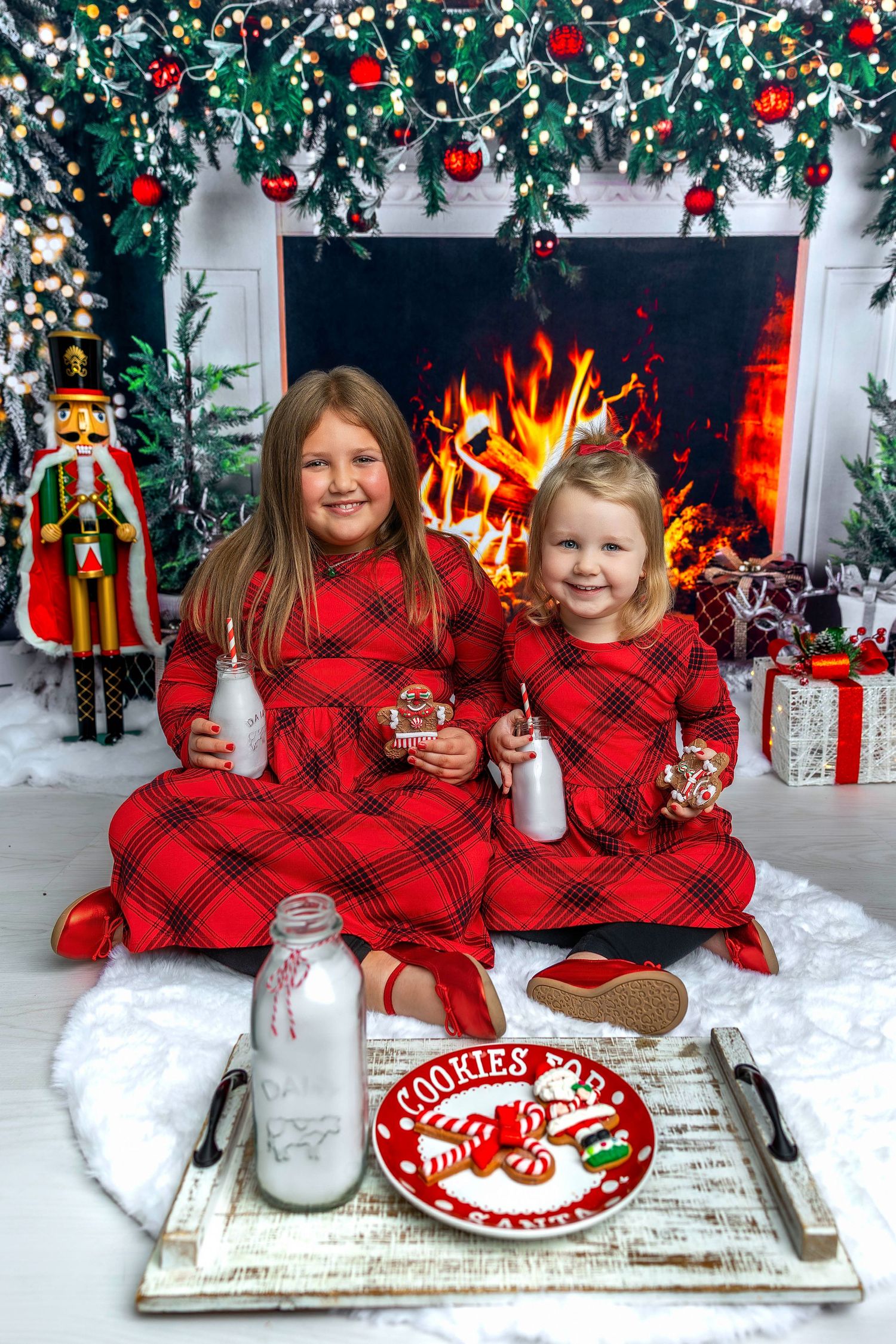 A festive holiday scene with matching red plaid outfits against a white and green Christmas backdrop with presents and decor.