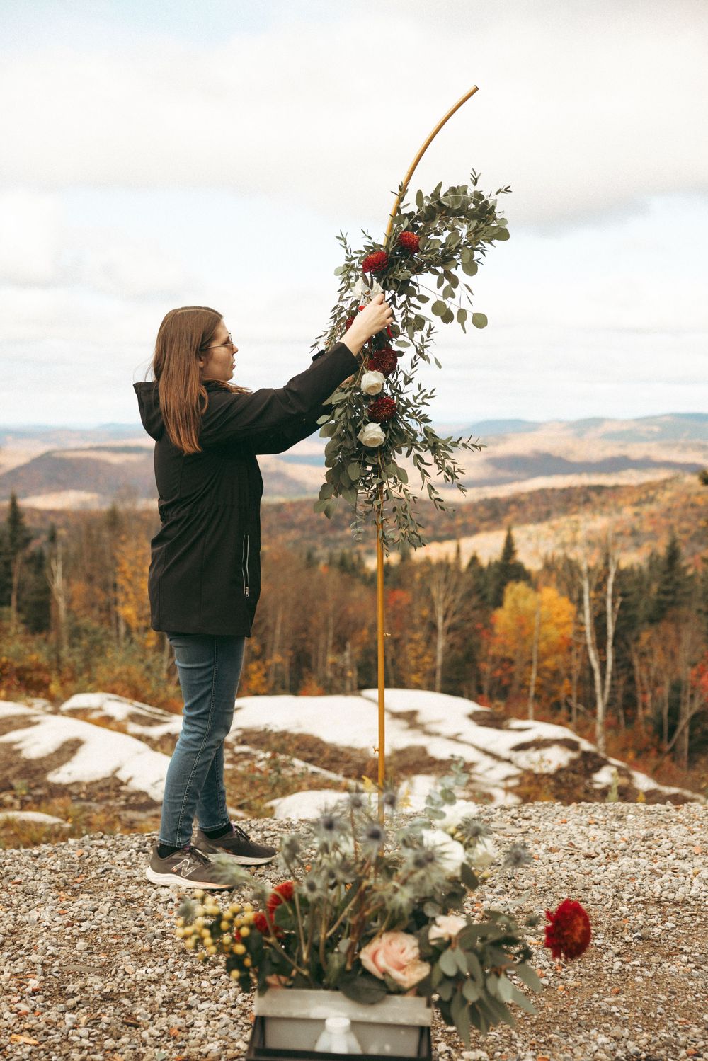 Person arranging floral decorations on a mountaintop with snow patches and autumn trees in the background.
