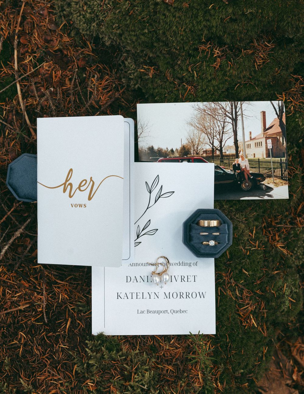 Wedding stationery flat lay featuring invitation suite, rings, and 'her' vows book on dark background.