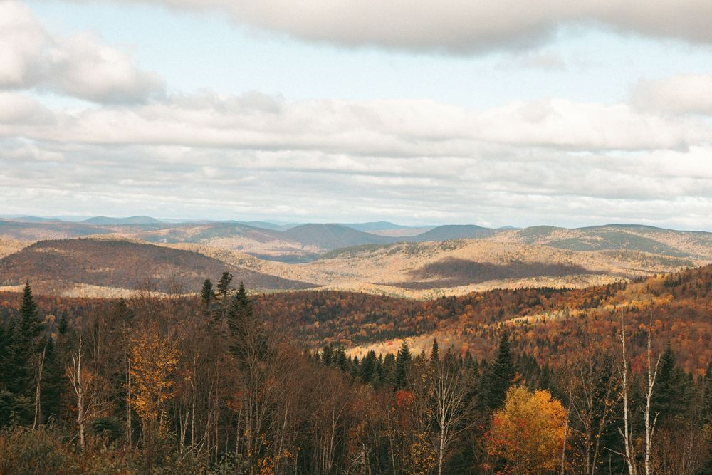 Panoramic autumn landscape showing rolling mountains and valleys with colorful fall foliage under cloudy skies.