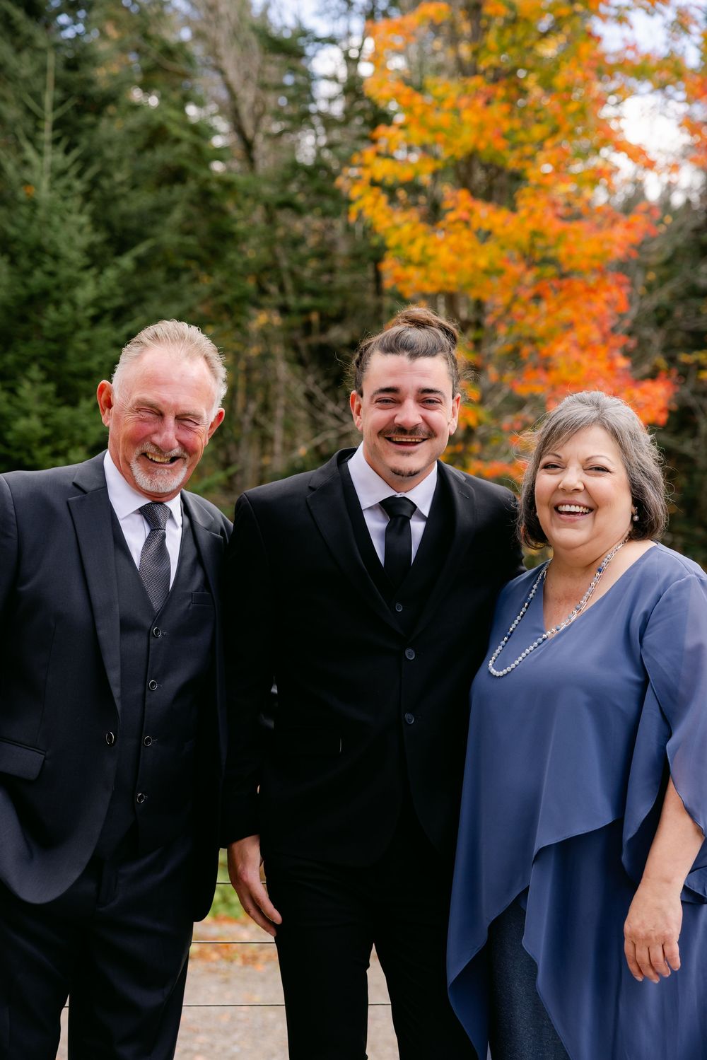 Group photo outdoors with autumn foliage featuring people in formal attire smiling and laughing together.