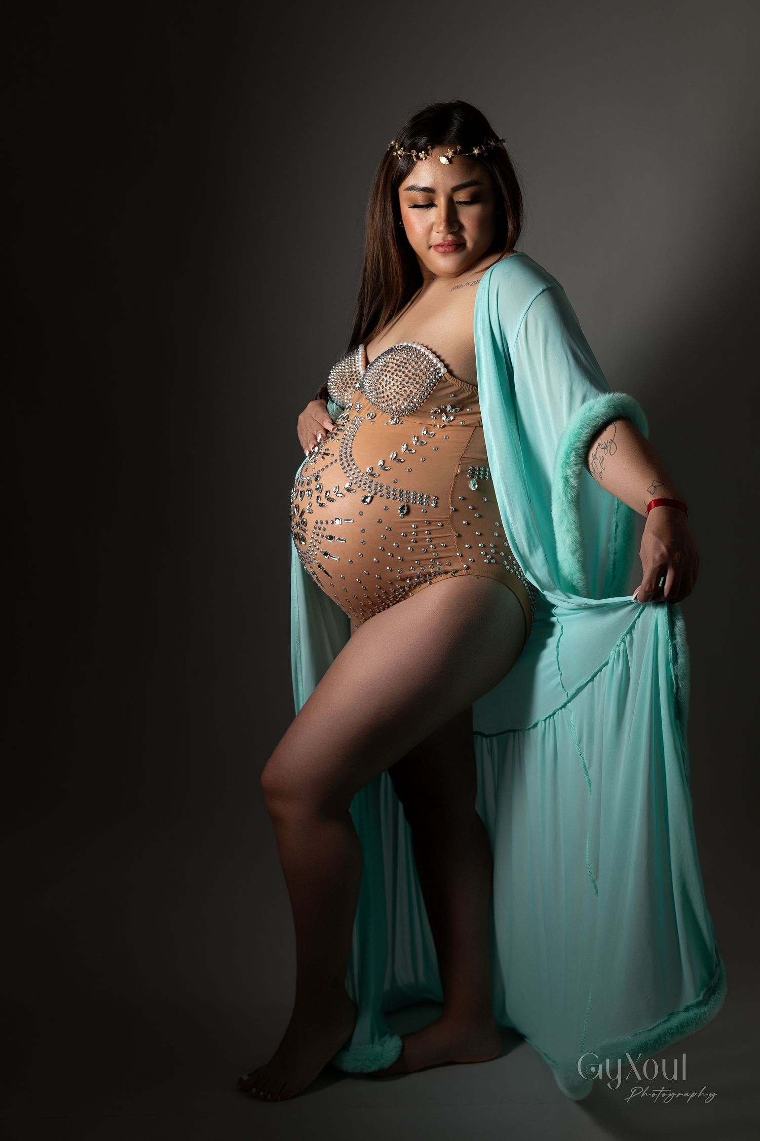 Dancer in turquoise robe and beaded bodysuit poses elegantly against dark background in dramatic studio lighting.