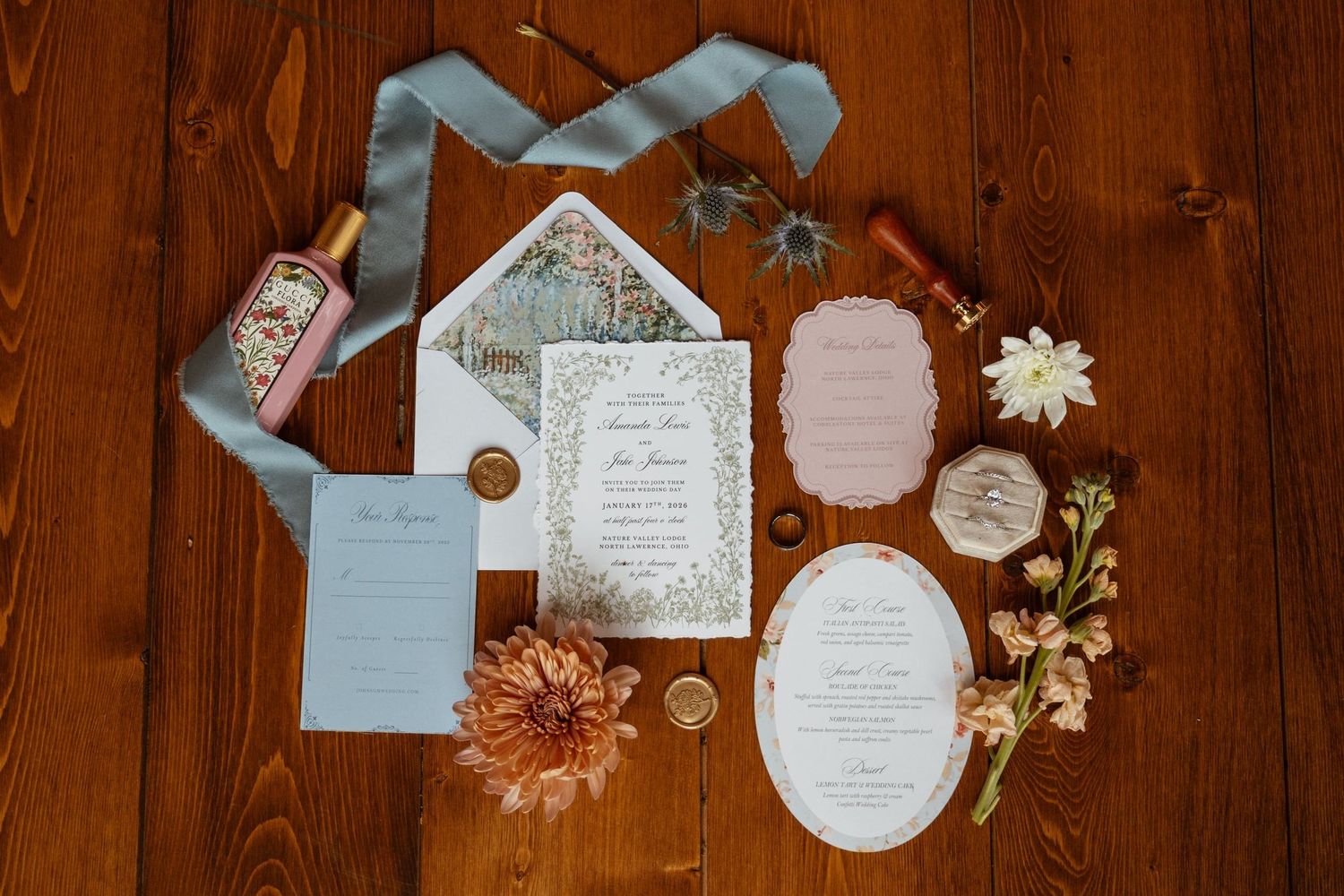 Wedding invitation suite with blue and floral details laid out on wooden surface with fresh flowers.