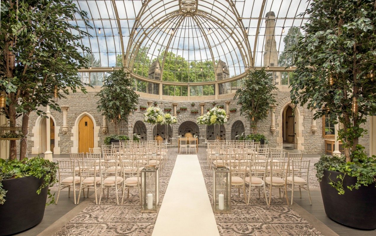 Elegant glass-roofed conservatory set up for a wedding ceremony with white aisle and wooden chairs.