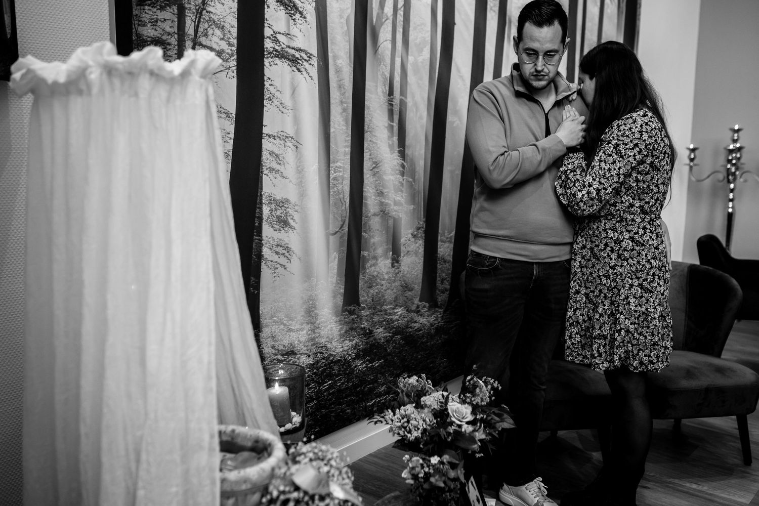 A black and white photo showing a wedding dress hanging while two figures interact in the background.