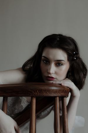 Moody portrait with dark hair and red lipstick leaning on wooden chair in dramatic lighting.