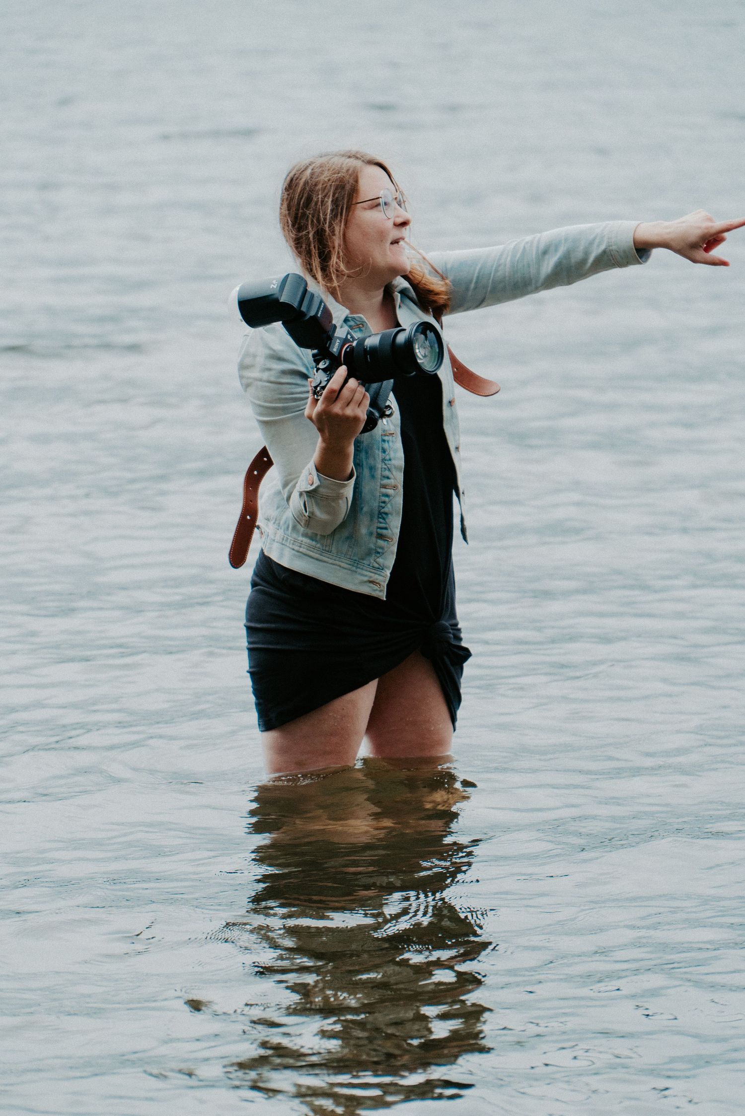 A person stands in shallow water wearing a denim jacket and black dress while pointing towards the horizon.