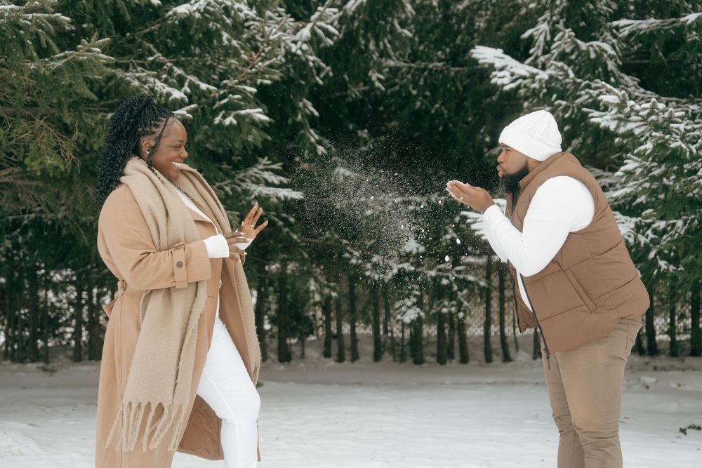A couple shares a romantic moment in a snowy winter forest setting wearing cozy neutral-toned outfits.