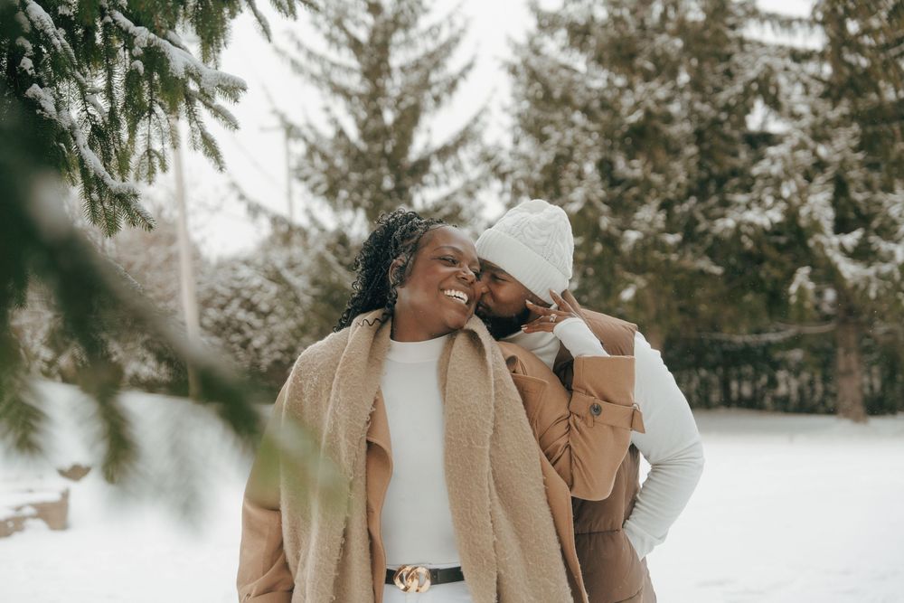 A couple shares a tender moment outdoors in winter, wearing cozy sweaters against a snowy evergreen backdrop.