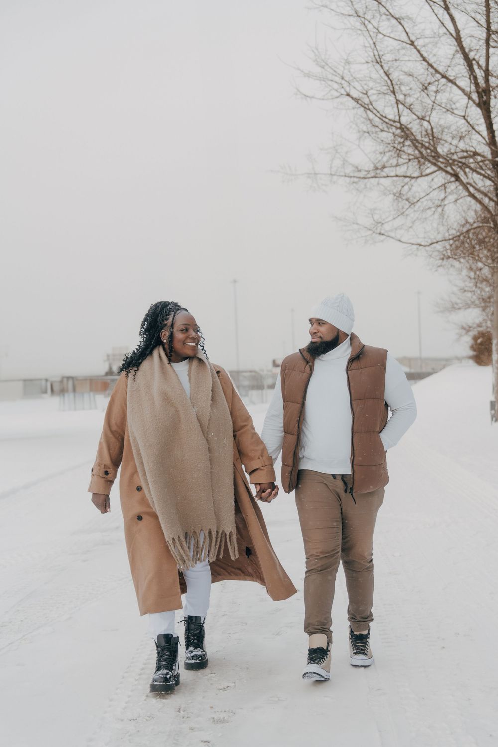 A couple in matching tan winter coats walks hand in hand through a snowy winter landscape.