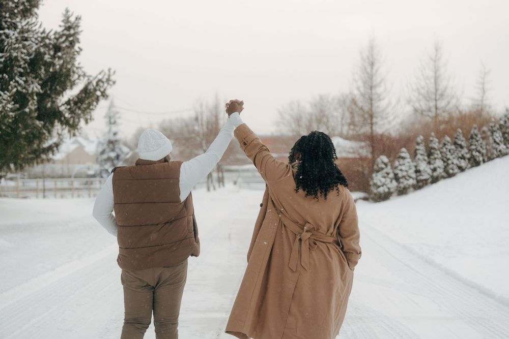 Two people in winter coats hold hands while walking down a snowy path lined with evergreen trees.
