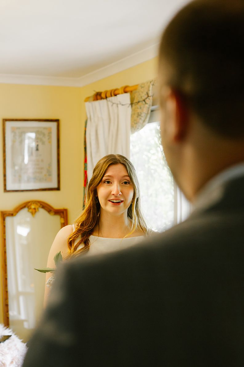 A bride wearing a white dress smiles warmly during her wedding day in a yellow-walled room with natural lighting.
