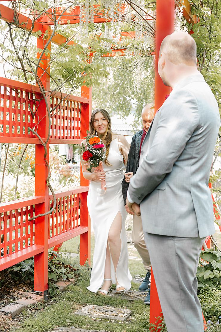 Wedding ceremony takes place in garden setting with bright red Asian-inspired lattice structure and lanterns overhead.