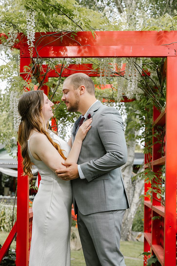 Romantic wedding couple shares intimate moments under red Japanese garden archway in outdoor setting.