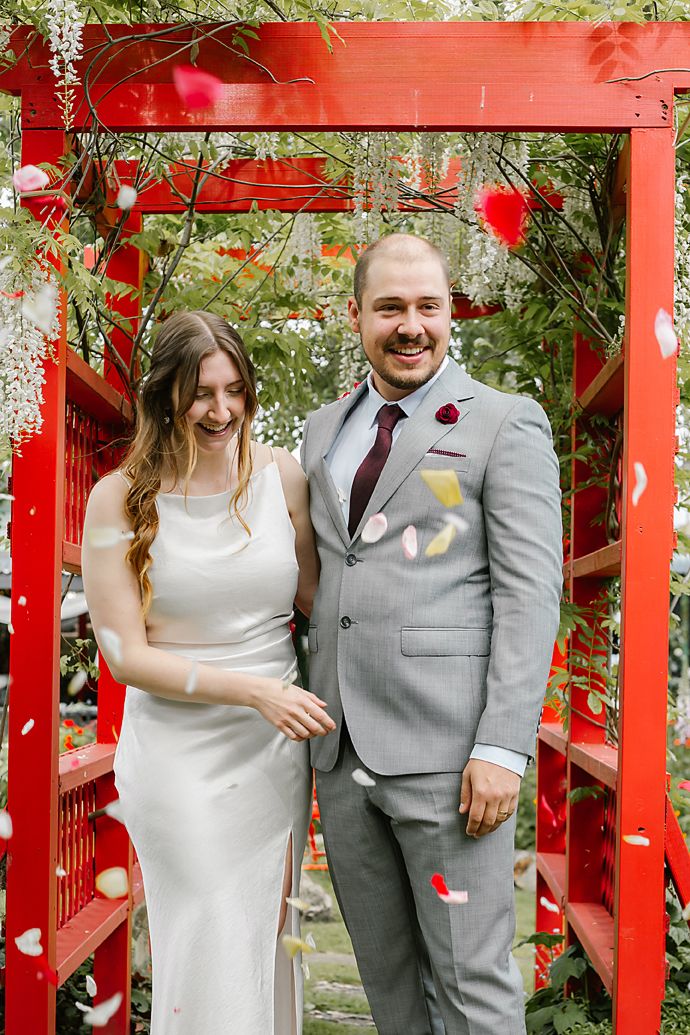 A couple poses together in front of a bright red garden trellis with greenery in the background.