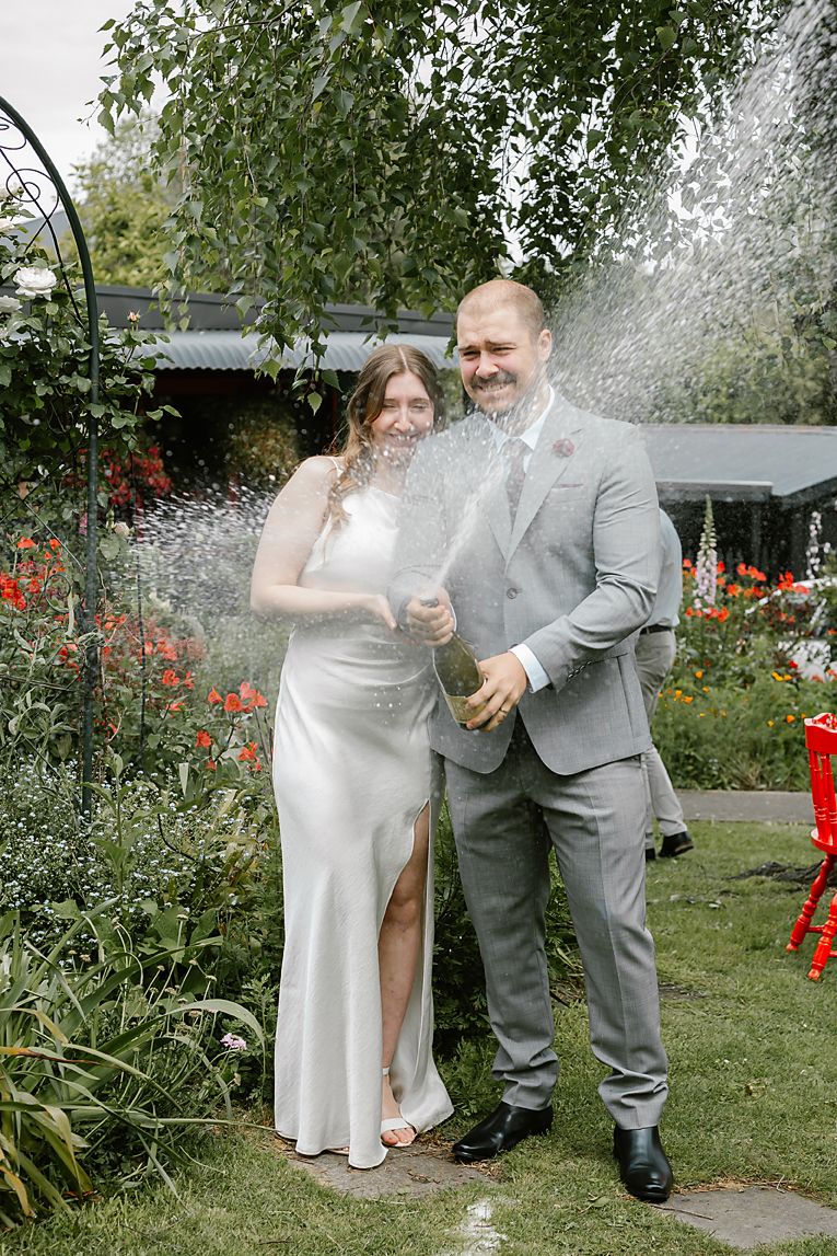 A couple in wedding attire celebrates with champagne spray in a garden setting with red flowers in the background.