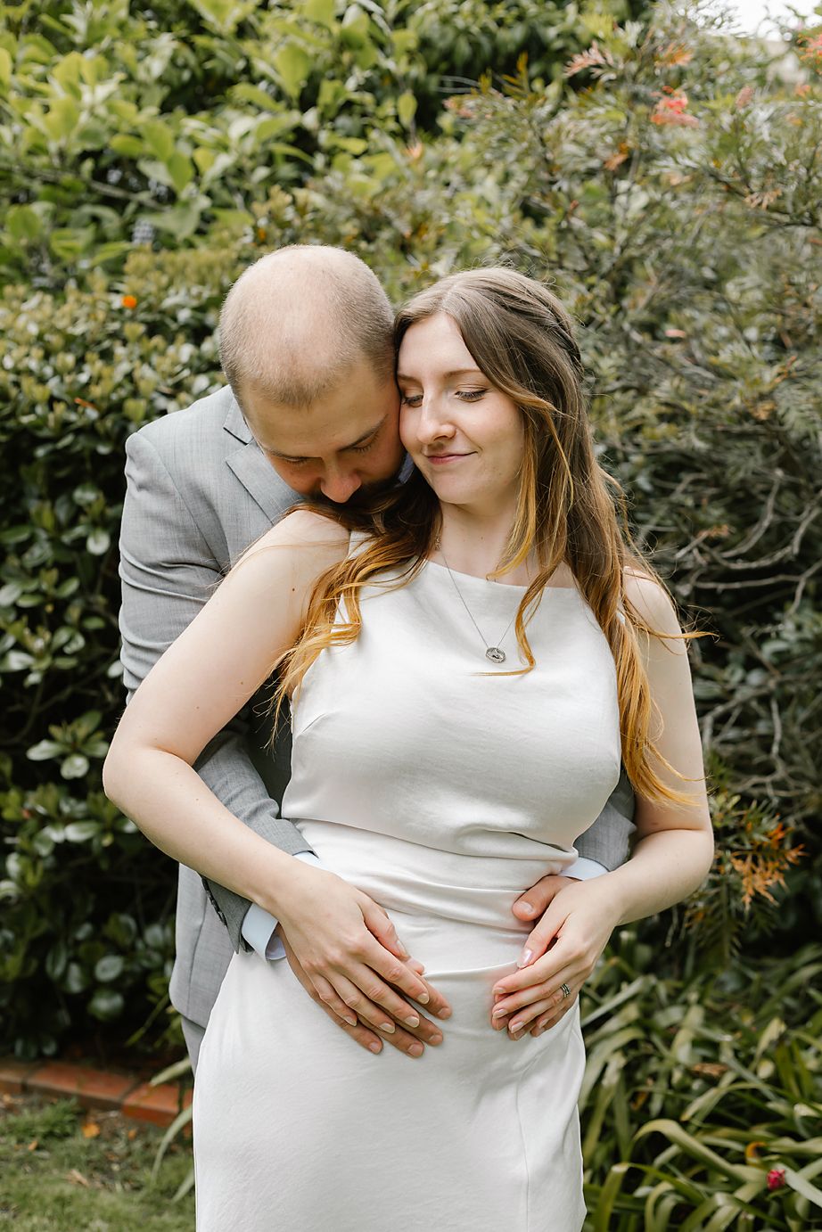 A romantic garden maternity photo shows a couple embracing in front of lush greenery on a sunny day.