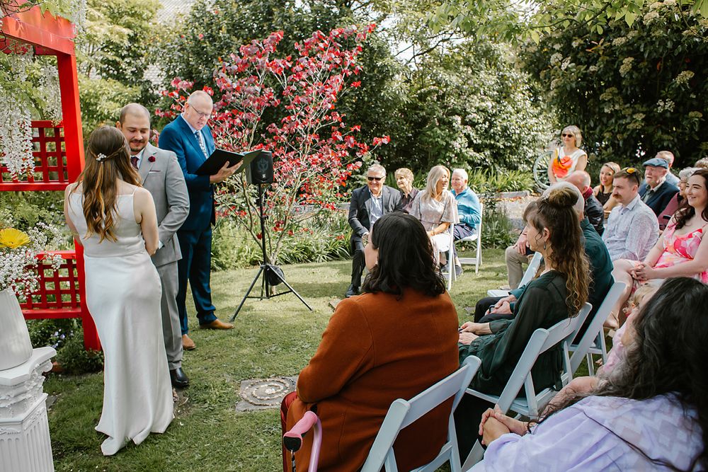A springtime outdoor wedding ceremony takes place in a garden with pink cherry blossoms blooming in the background.