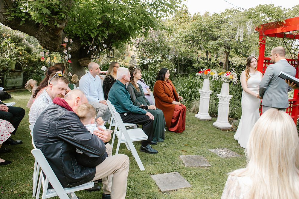 Guests gather in an outdoor garden setting during a wedding ceremony with white chairs arranged on the lawn.