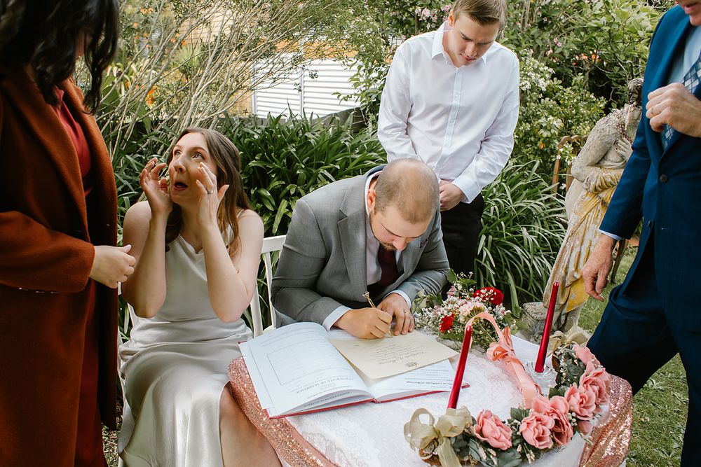 Wedding guests take turns signing the guest book at an outdoor garden ceremony surrounded by greenery and flowers.