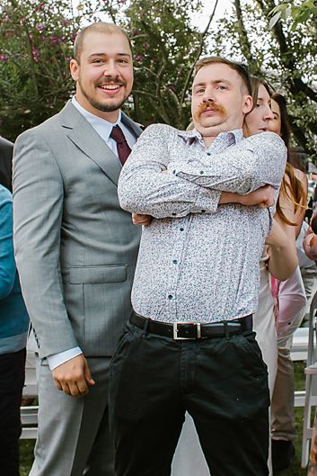 Two people in formal attire share a playful moment at an outdoor event with trees in the background.