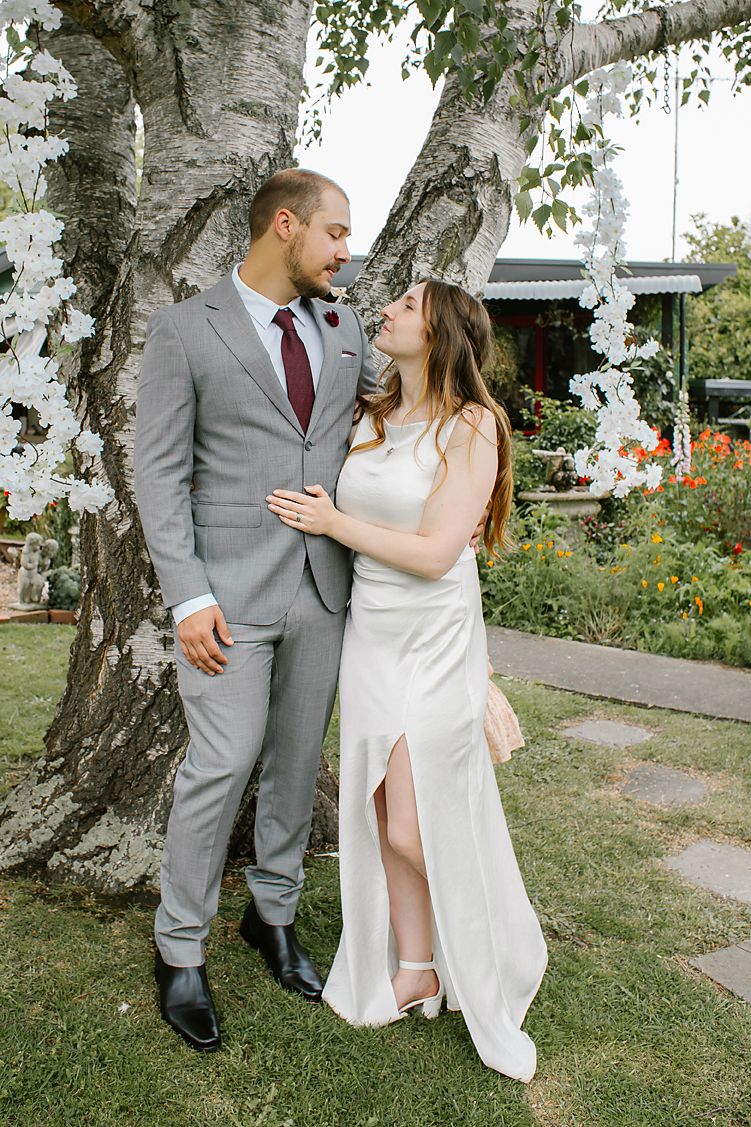 A couple in wedding attire dances playfully among birch trees in a garden setting.