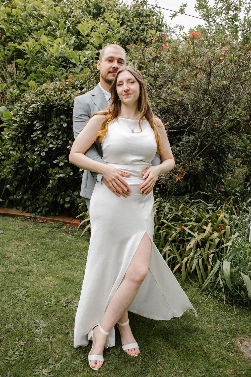 A couple poses together outdoors on grass with green foliage in the background.