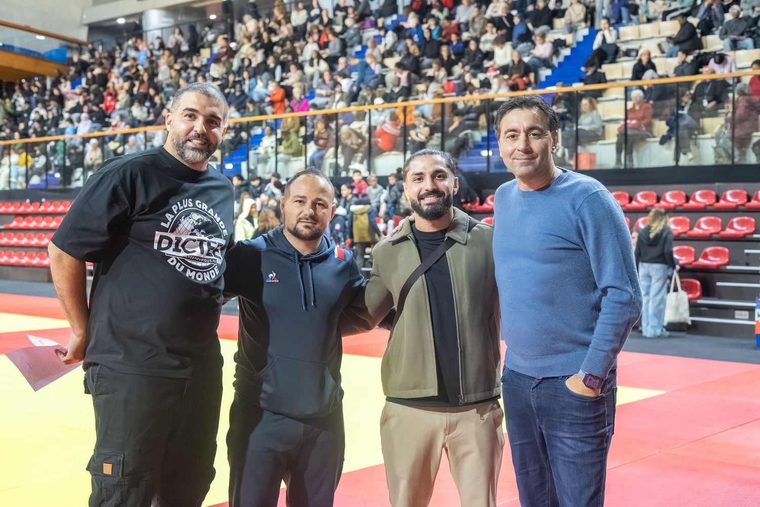 A group of people standing together at a martial arts competition venue with red mats visible in the background.