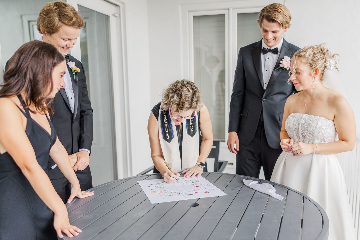Wedding ceremony participants gather around a gray table on a porch to sign documents.