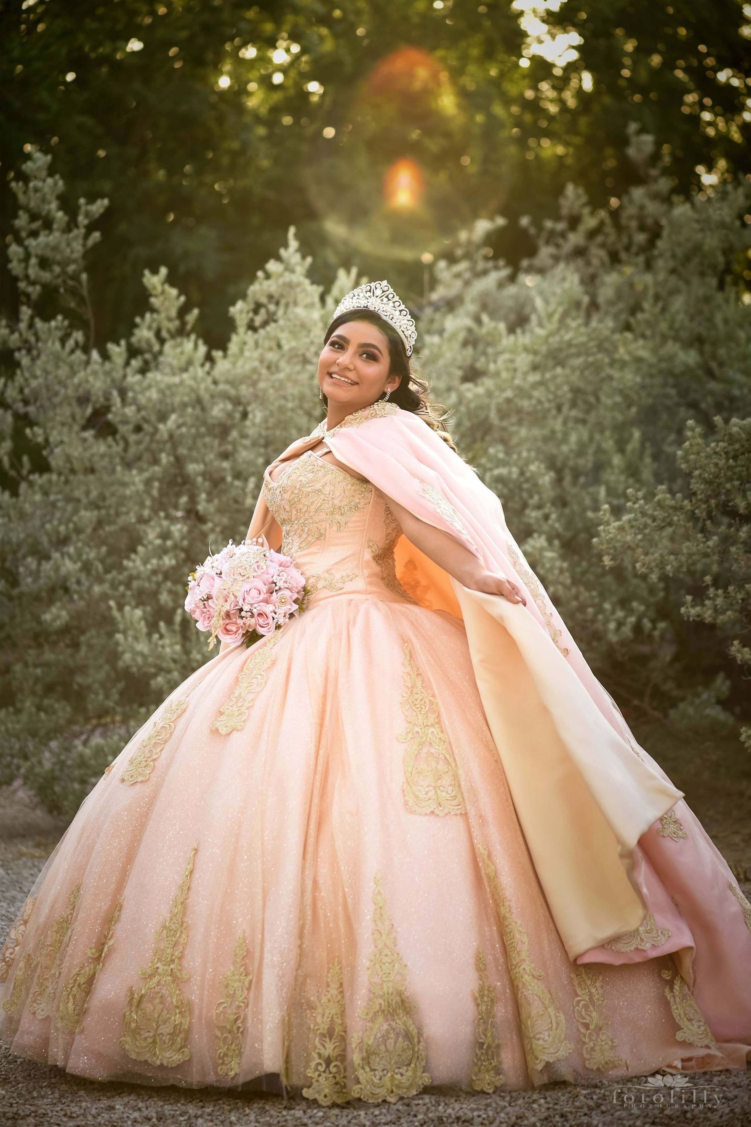 A stunning pink and gold quinceanera ball gown with a full tulle skirt photographed in a sunlit outdoor setting.