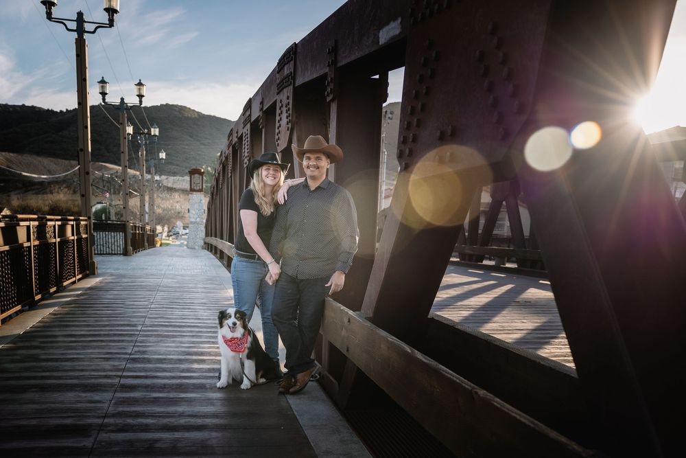A couple and their dog stand on a wooden boardwalk at sunset with mountains and lampposts in the background.
