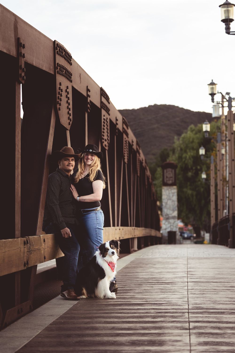 A couple and their dog pose together on a rustic wooden boardwalk with mountains in the background.