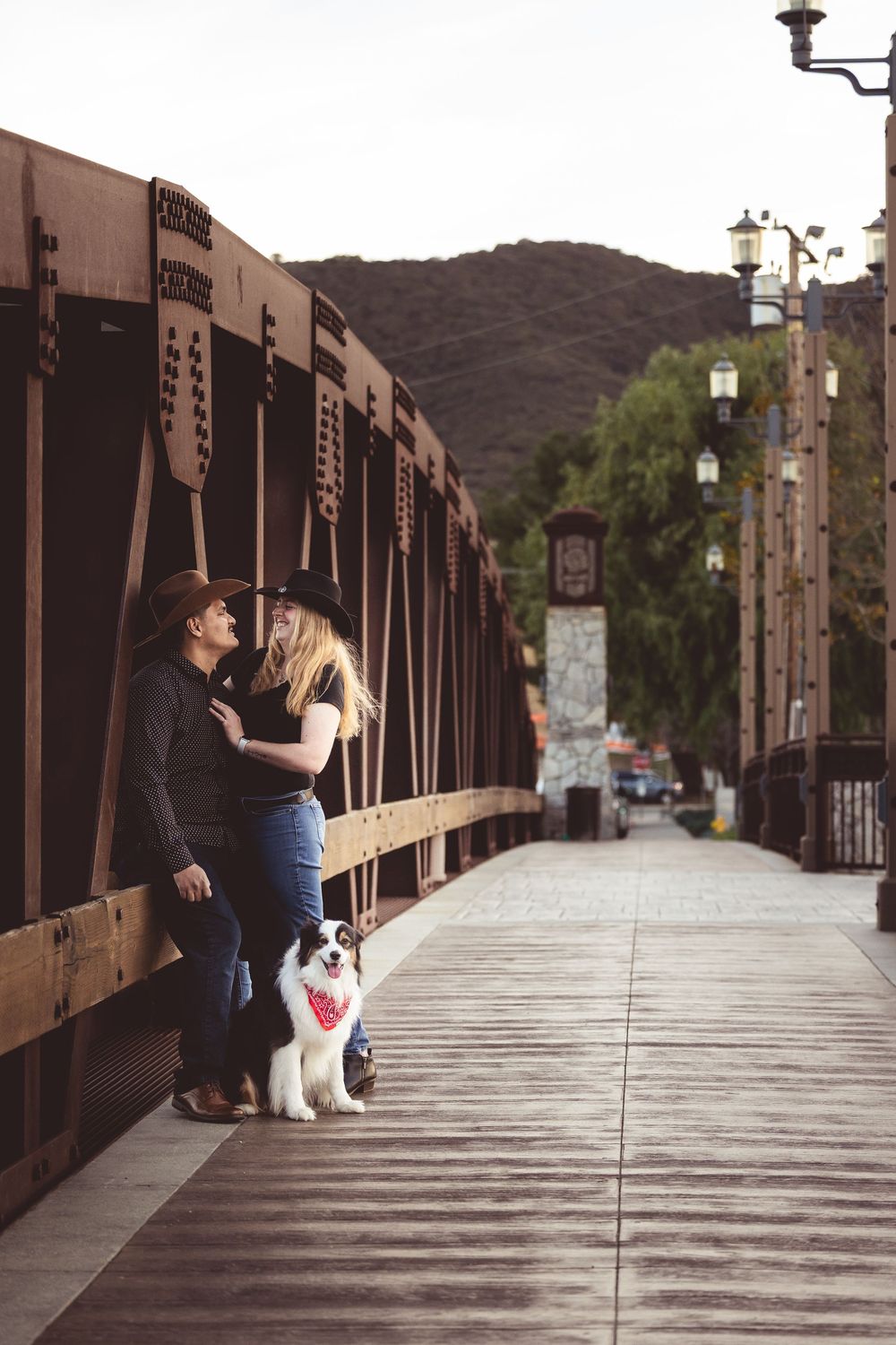 A couple and their white dog share a tender moment on a rustic wooden bridge walkway at sunset.
