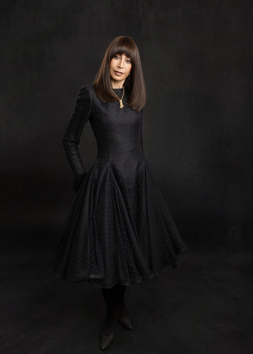 A person in a long-sleeved black dress with full skirt poses elegantly against a dark background in a studio setting.