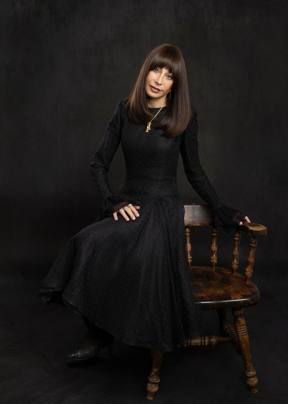 A model in a black dress poses elegantly on a wooden chair against a dark background in a dramatic studio setting.