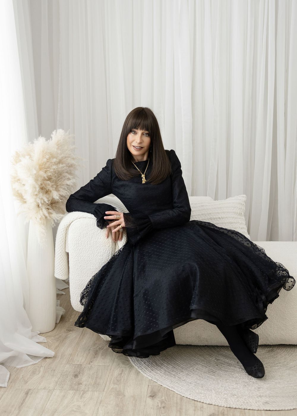 A model in a black dress reclines on a white sofa with pampas grass decor in a bright, minimalist setting.