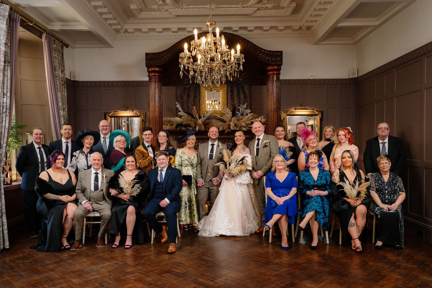 Elegant wedding party group photo in a grand room with ornate chandelier and classic decor. Ellingham Hall