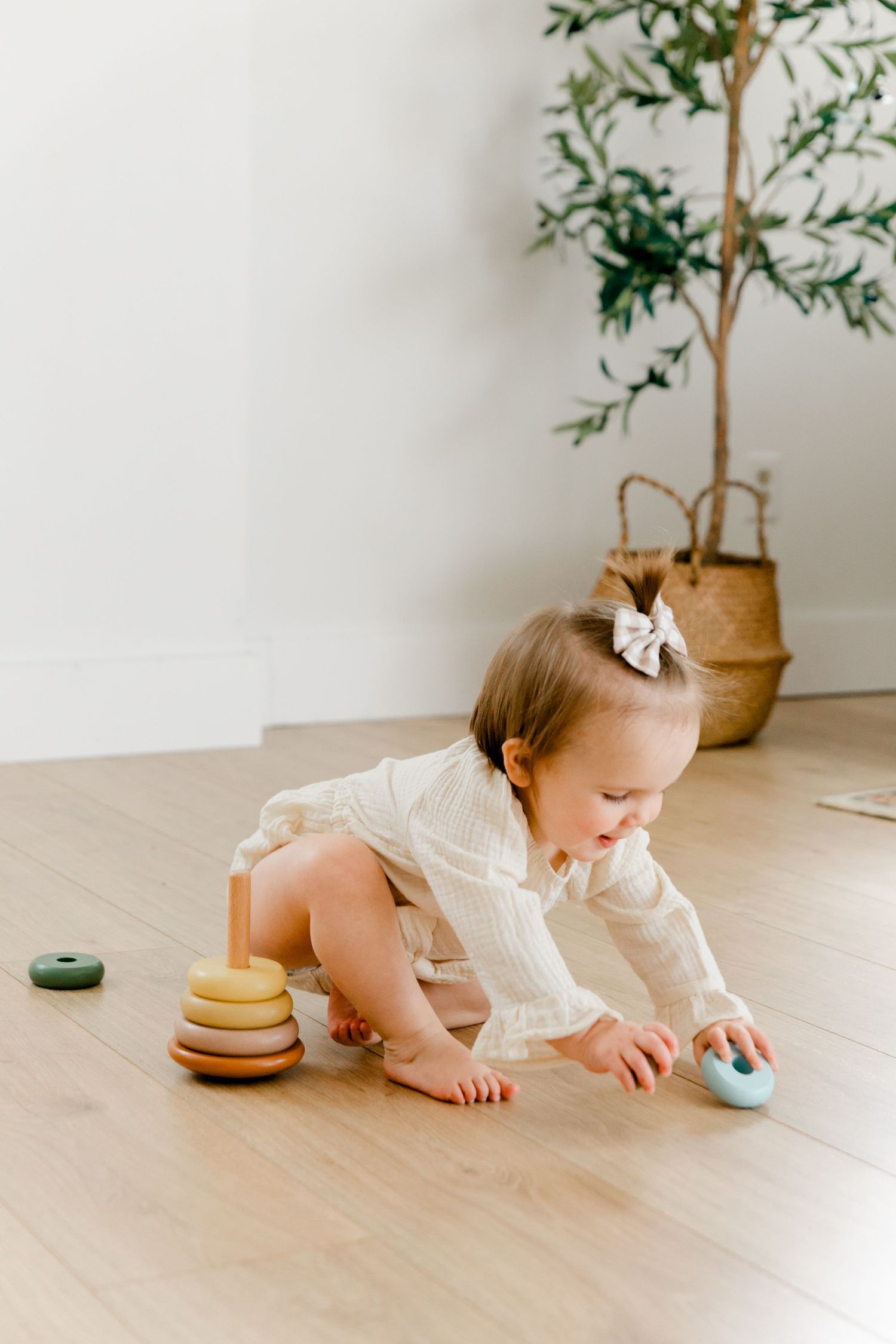 Baby girl in cream outfit reaching for toy on hardwood floor with plant in background.
