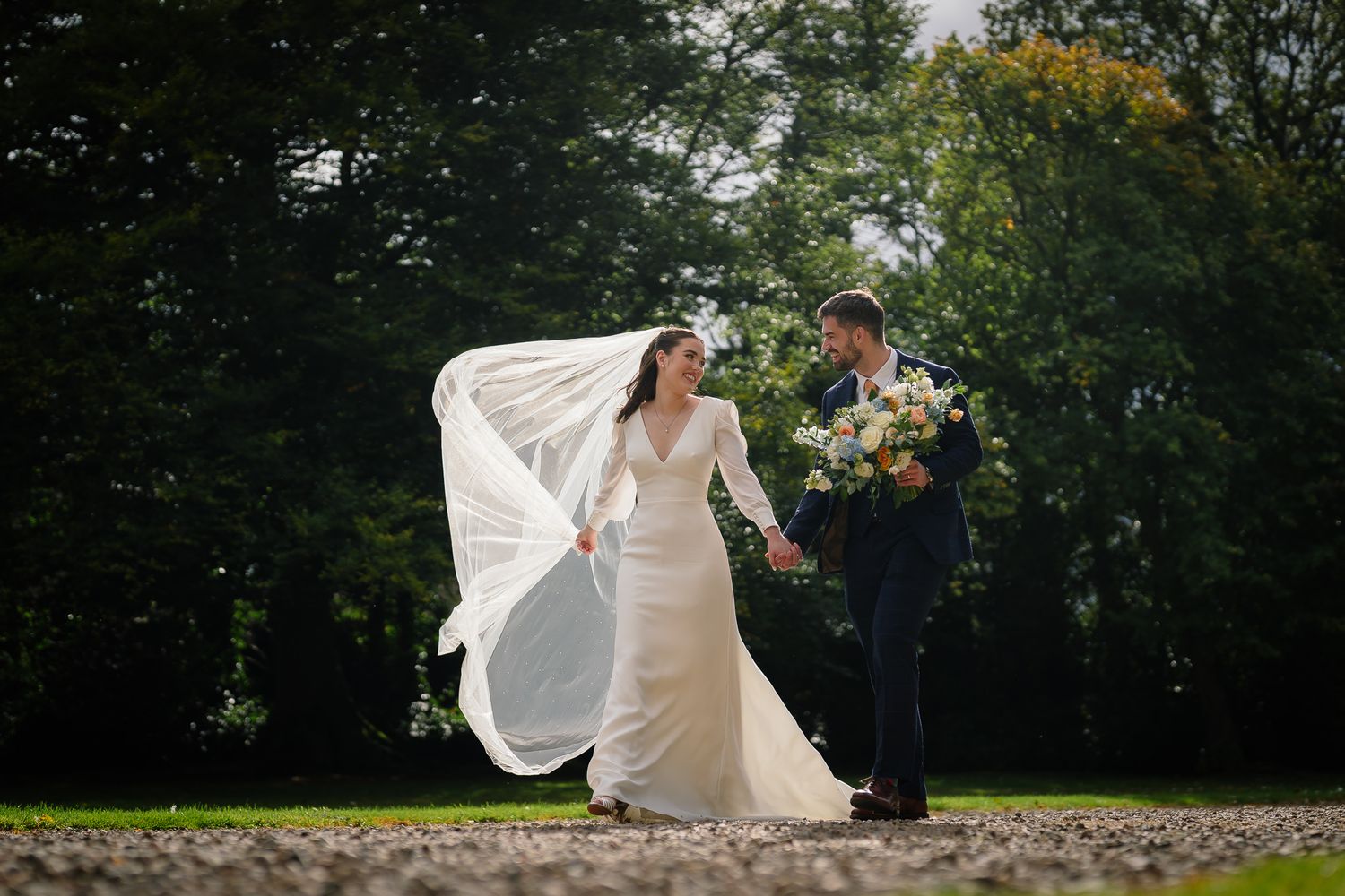 Wedding dress flowing in breeze against backdrop of green garden foliage. Ellingham Hall