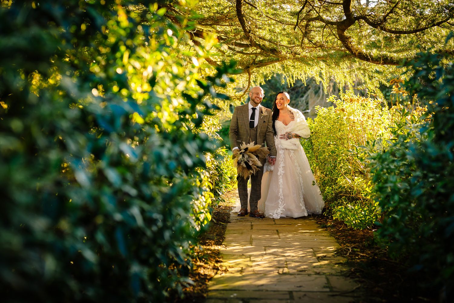 Wedding couple walking through lush garden path with golden sunlight filtering through trees. Ellingham Hall