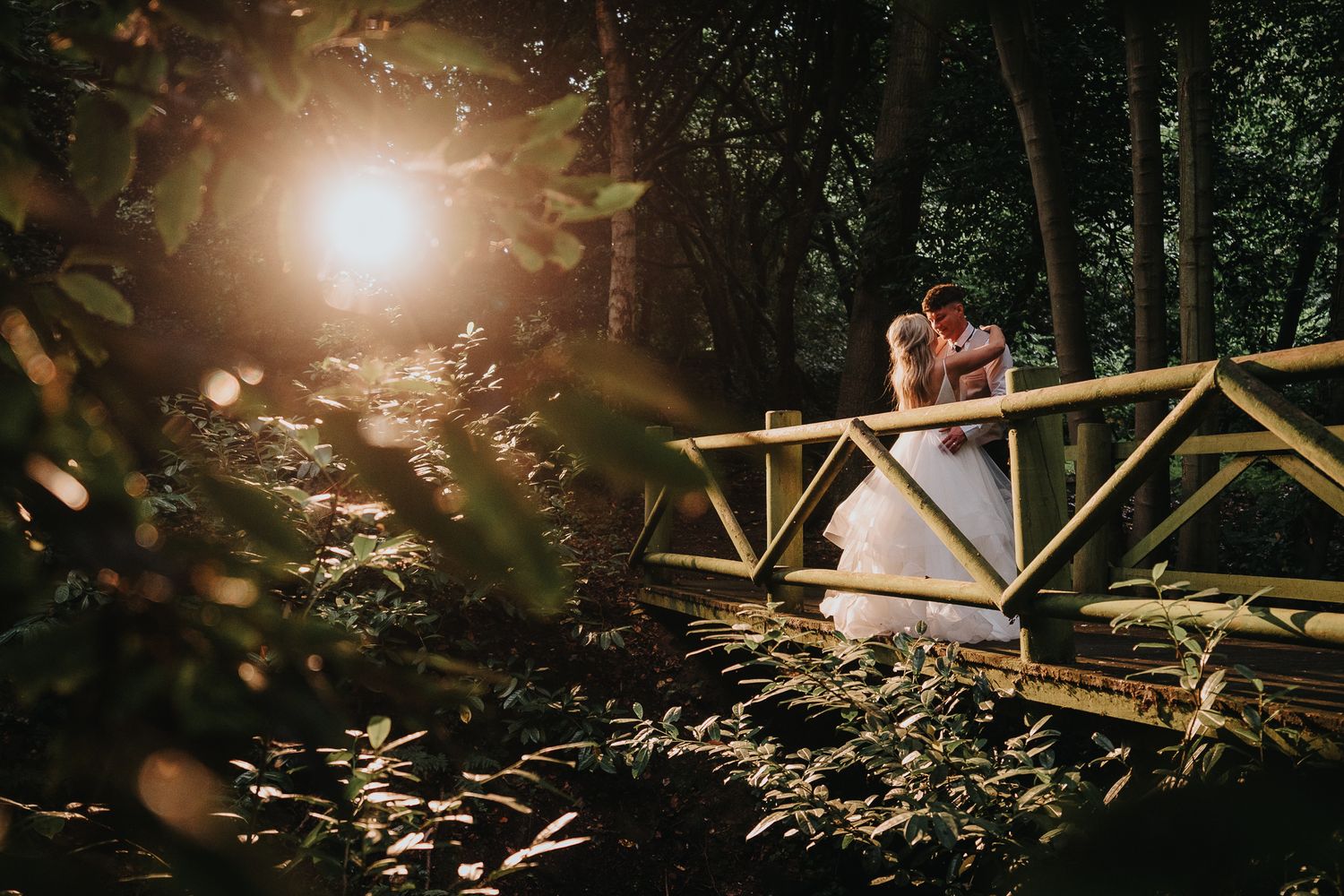 Wedding couple silhouetted against golden sunlight on a rustic wooden bridge in a forest setting. Ellingham Hall