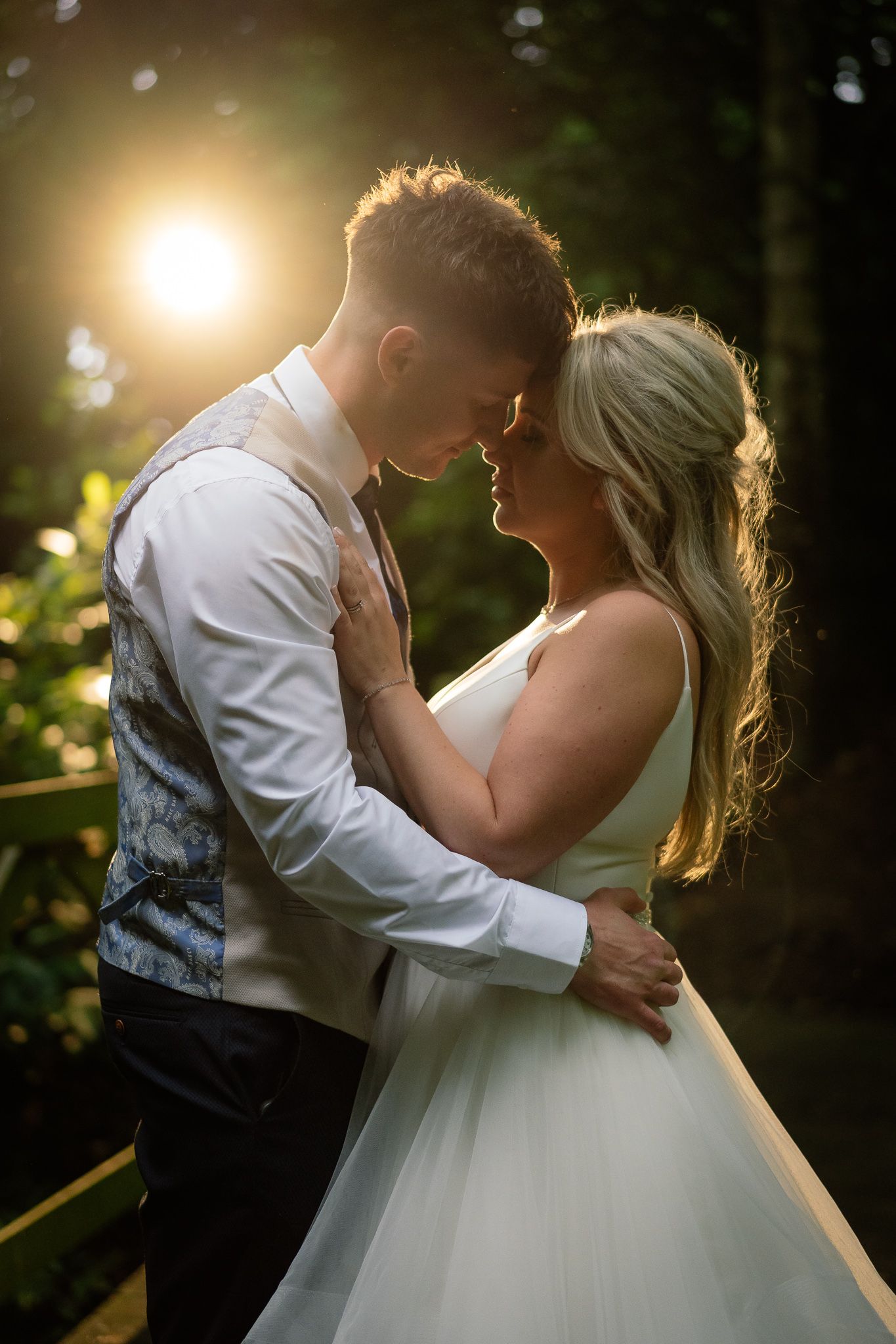 Romantic sunset portrait of a couple embracing in formal wedding attire with golden backlighting. Ellingham Hall
