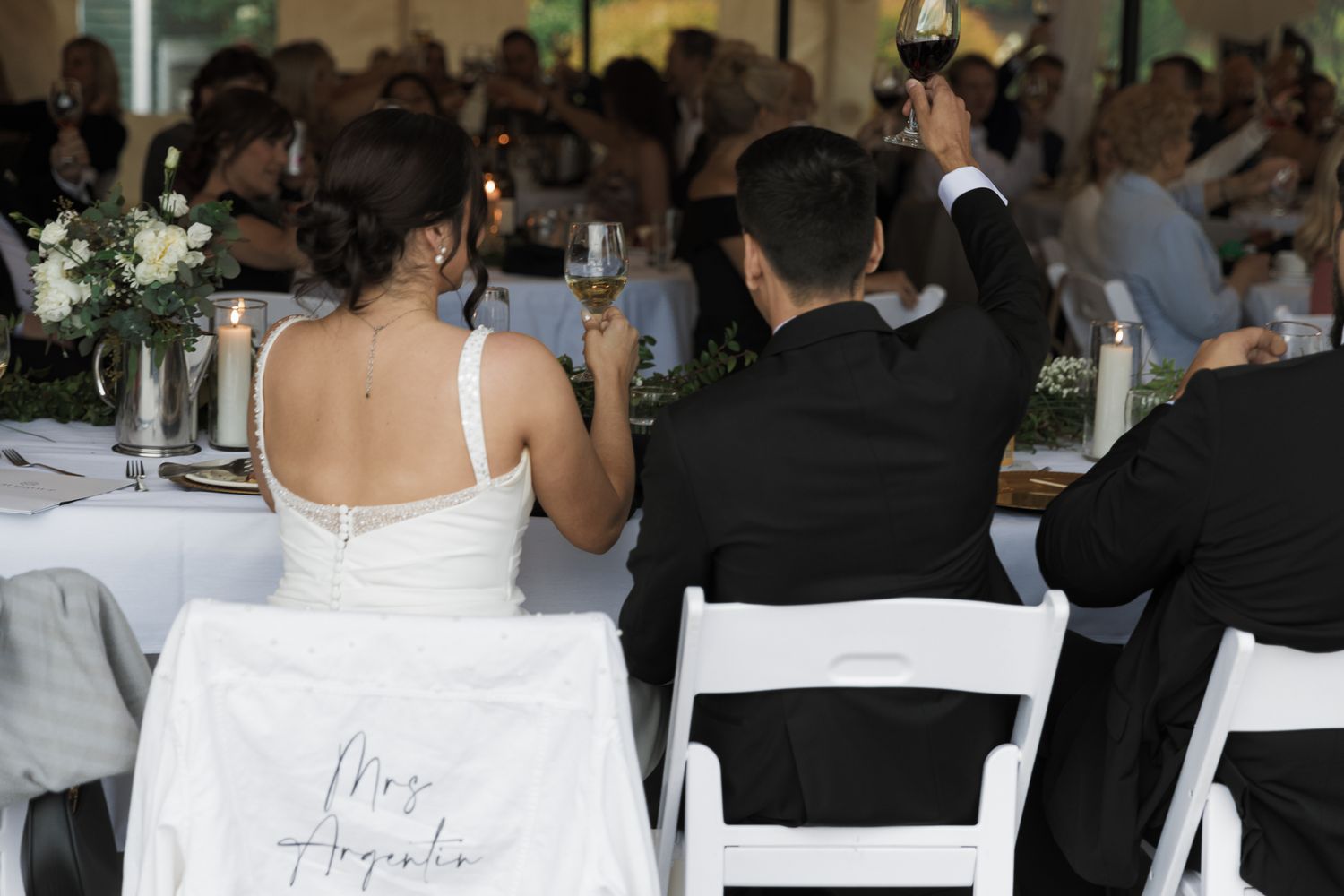Bride and groom seated at wedding reception head table with Mrs. sign on bride's chair during toasts.