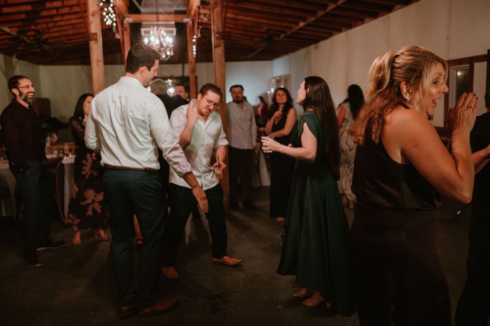 Wedding guests dance and socialize at an indoor reception with exposed brick walls and wooden beams.