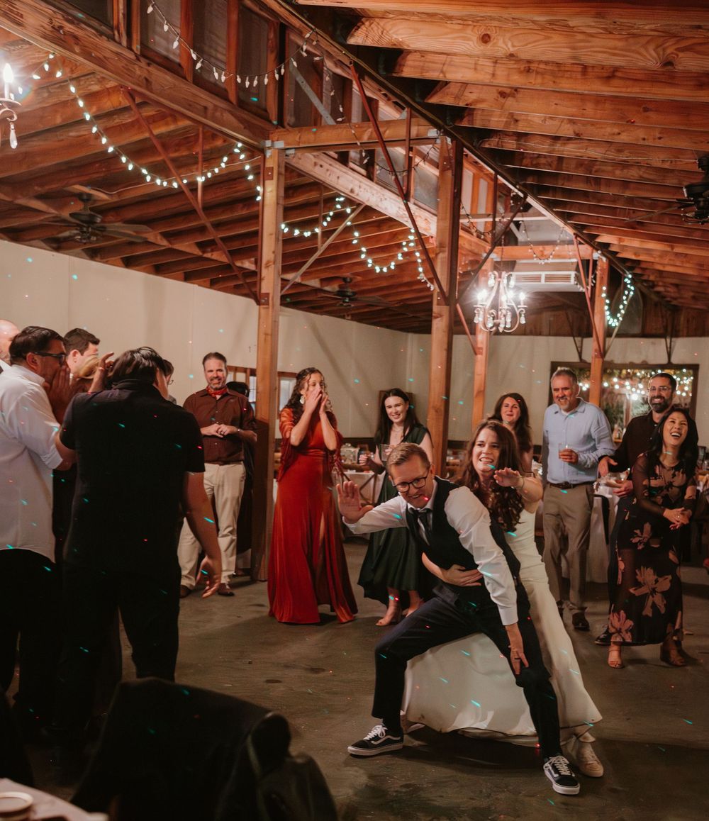 Wedding reception guests dancing and celebrating under rustic wooden beams with string lights.