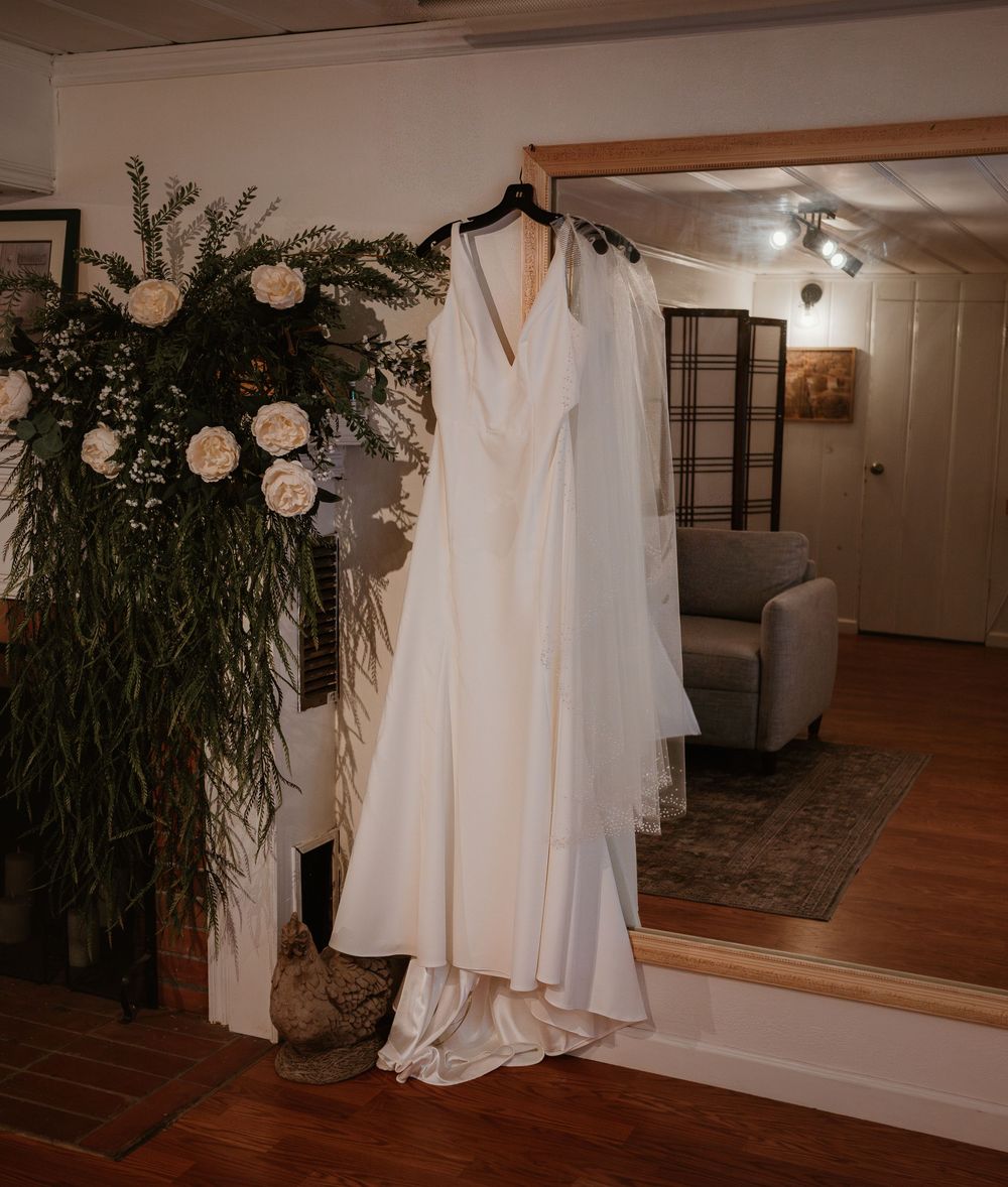 Elegant white wedding dress hanging in a rustic venue with greenery and floral decorations.