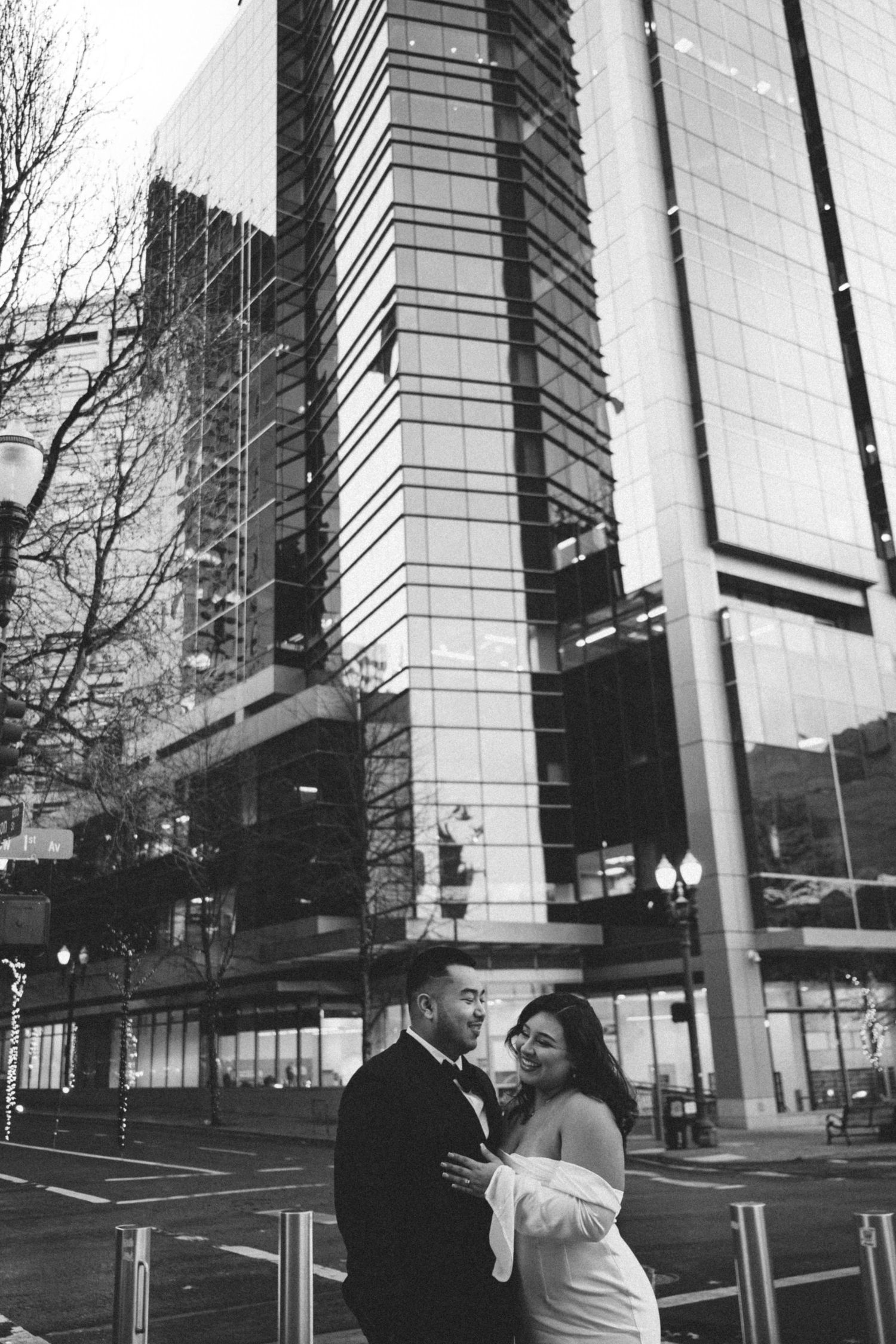 A couple shares a romantic moment in front of a modern glass office building in black and white photography.